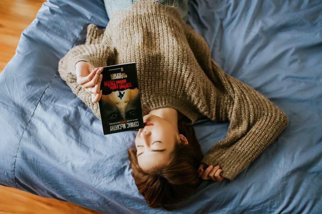 woman in sweater holding book while lying on bed